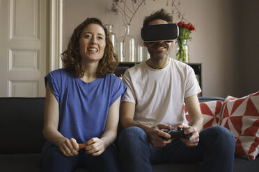 Smiling woman sitting by man playing on virtual reality headset in living room at home - FSIF03121