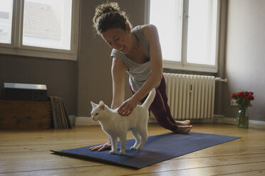 Smiling woman stroking white cat while practicing yoga at home - FSIF03123