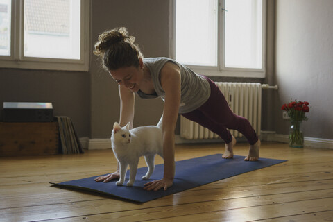 Smiling woman practicing plank position over white cat on exercise mat at home stock photo