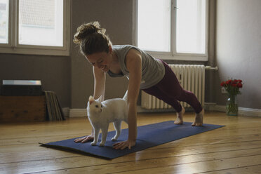 Smiling woman practicing plank position over white cat on exercise mat at home - FSIF03127