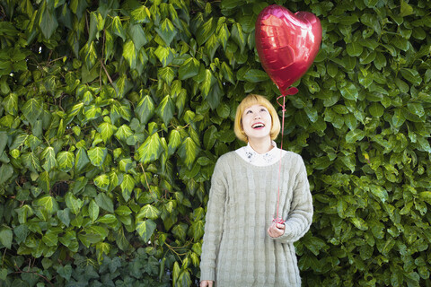 Lächelnde junge Frau, die einen herzförmigen Ballon hält, während sie vor Pflanzen steht, lizenzfreies Stockfoto