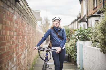 Porträt einer jungen Frau, die mit ihrem Fahrrad auf einem Fußweg an einer Mauer spazieren geht - FSIF03159