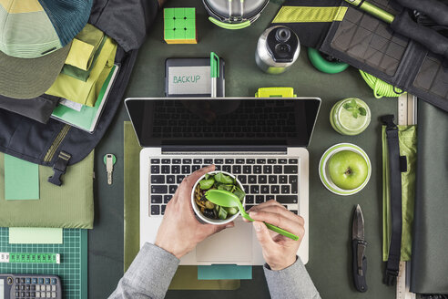 Draufsicht auf die Hände eines Mannes, der Salat hält, während er Wanderausrüstung und Laptop vorbereitet - CUF23150