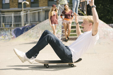 Young male skateboarder sitting on skateboard on the move in city skatepark - CUF23158