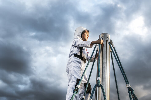 Junge im Astronautenkostüm blickt von einem Klettergerüst in den dramatischen Himmel - CUF23192