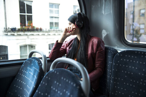 Junge Frau sitzt im Bus, trägt Kopfhörer und schaut aus dem Fenster - CUF23235