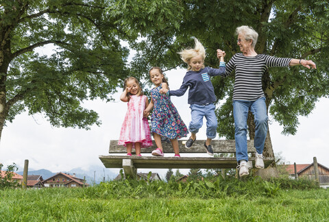 Großmutter und Enkelkinder springen in der Luft von einer Parkbank, Füssen, Bayern, Deutschland, lizenzfreies Stockfoto