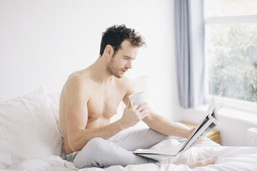 Junger Mann liegt im Bett, trinkt Kaffee und liest Zeitung - CUF23314