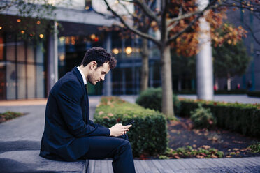 Geschäftsmann sitzt vor seinem Büro und schreibt eine SMS auf seinem Smartphone - CUF23318