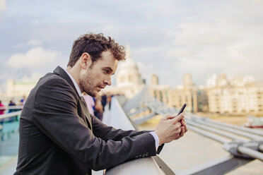 Businessman reading smartphone texts whilst leaning on millennium bridge, London, UK - CUF23334