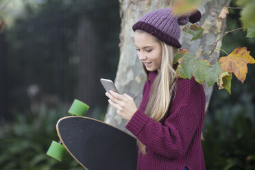 Smiling teenage girl looking at cell phone holding skateboard - ZEF15595