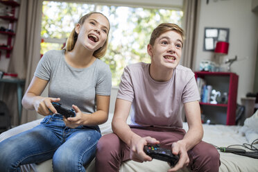 Happy teenage girl and boy sitting on bed playing video game - ZEF15642