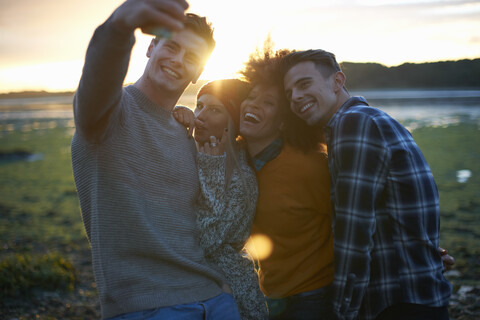 Vier junge Erwachsene machen ein Smartphone-Selfie bei Sonnenuntergang am Meer, lizenzfreies Stockfoto