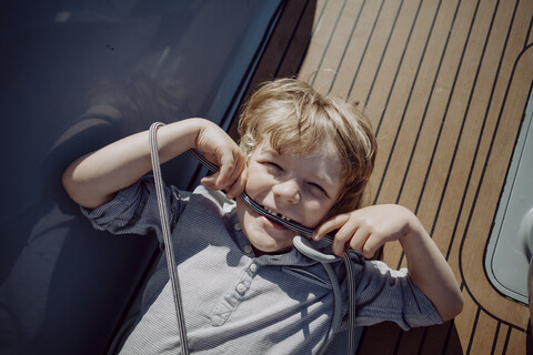 Verspielter Junge an Deck eines Segelboots mit einem Seil im Mund, lizenzfreies Stockfoto
