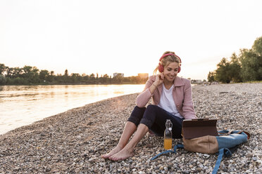 Young blond woman with earphones and tablet on riverside in the evening - UUF14066