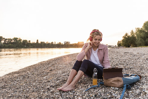 Junge blonde Frau mit Kopfhörern und Tablet am Flussufer am Abend - UUF14066