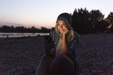 Junge Frau benutzt ihr Smartphone abends am Flussufer - UUF14070