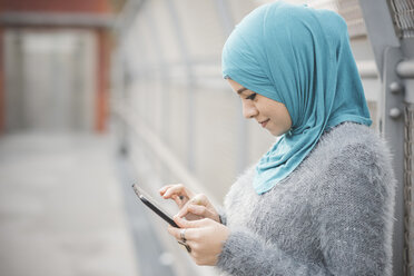 Junge Frau mit türkisfarbenem Hidschab, die auf einer Fußgängerbrücke ein digitales Tablet benutzt - CUF23609