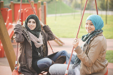 Portrait of two young women sitting on playground swings - CUF23625