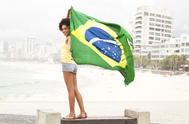 Porträt einer jungen Frau, die auf einer Parkbank die brasilianische Flagge hochhält, Strand von Ipanema, Rio De Janeiro, Brasilien - CUF23655