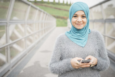 Porträt einer jungen Frau mit türkisfarbenem Hidschab, die auf einer Fußgängerbrücke ihr Smartphone benutzt - CUF23661