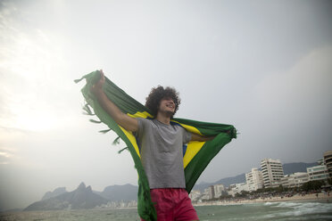 Lächelnder junger Mann hält die brasilianische Flagge am Strand von Ipanema hoch, Rio De Janeiro, Brasilien - CUF23683