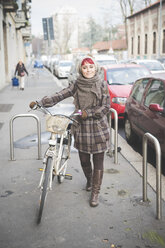 Porträt einer jungen Frau in Winterkleidung, die mit dem Fahrrad die Straße entlanggeht - CUF23731