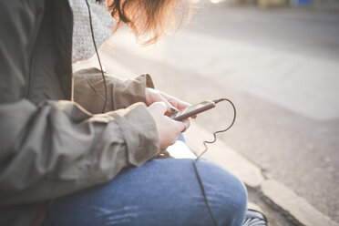 Cropped shot of young woman sitting on sidewalk reading smartphone texts - CUF23760