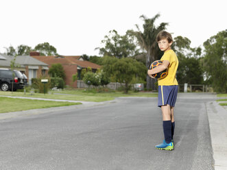 Porträt eines mürrischen Jungen, der auf einer Vorstadtstraße einen Fußball hält - CUF24109