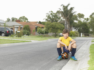 Porträt eines auf einem Fußball sitzenden Jungen auf einer Vorstadtstraße - CUF24111