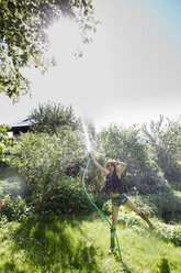 Vorderansicht einer reifen Frau im Garten, die auf einem Bein steht und mit einem Schlauch Wasser in die Luft spritzt - CUF24412