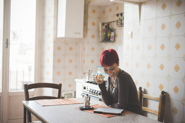 Junge Frau sitzt am Tisch, trinkt Kaffee und schaut auf ein digitales Tablet - CUF24493