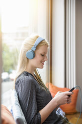 Young woman, wearing headphones, sitting in cafe, holding smartphone - CUF24533