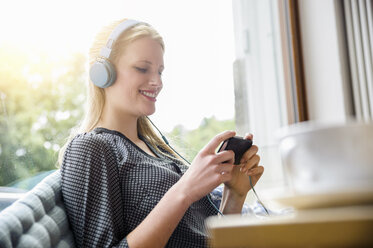 Young woman, wearing headphones, sitting in cafe, holding smartphone - CUF24534