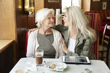 Mutter und Tochter sitzen zusammen in einem Café, der Arm der Tochter liegt um die Mutter, ein digitales Tablet liegt auf dem Tisch - CUF24663