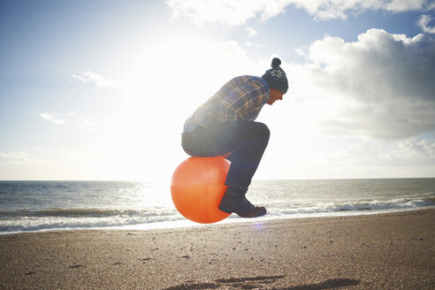 Mature man jumping mid air on inflatable hopper at beach - CUF24746