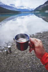 Cropped view of mans hand holding cup of tea by river, Banff, Alberta, Canada - ISF09352