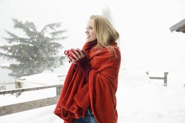 Junge Frau im verschneiten Nebel, eingewickelt in eine rote Decke, trinkt Kaffee - CUF25024