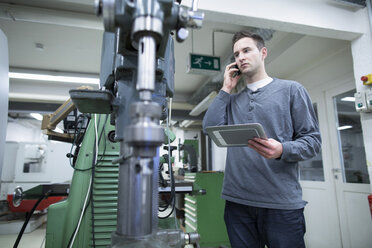 Junger männlicher Techniker, der in der Werkstatt mit seinem Smartphone chattet, während er ein digitales Tablet benutzt - CUF25212