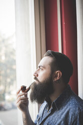 Young bearded man smoking pipe by window - CUF25615