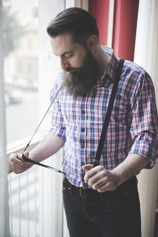 Junger bärtiger Mann, der am Fenster eine Zahnspange zieht, lizenzfreies Stockfoto