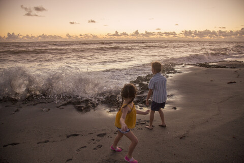 Junge und Schwester laufen vor den plätschernden Wellen am Strand bei Sonnenaufgang weg, Blowing Rocks Preserve, Jupiter Island, Florida, USA, lizenzfreies Stockfoto