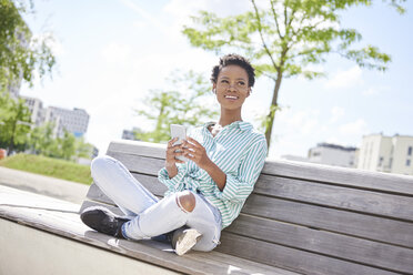 Portrait of smiling young woman with cell phone and earphones sitting on bench - ABIF00577