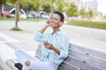 Porträt einer jungen Frau, die auf einer Bank sitzt und mit Handy und Kopfhörern Musik hört - ABIF00579