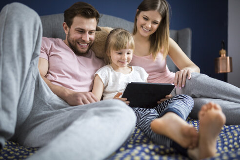 glückliche Familie, die auf dem Bett sitzt und mit ihrer kleinen Tochter etwas auf einem digitalen Tablet anschaut - AWF00020