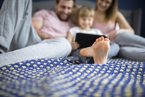 glückliche Familie, die auf dem Bett sitzt und mit ihrer kleinen Tochter etwas auf einem digitalen Tablet anschaut, lizenzfreies Stockfoto