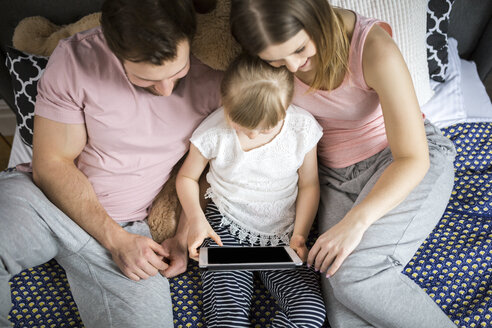 glückliche Familie, die auf dem Bett sitzt und mit ihrer kleinen Tochter etwas auf einem digitalen Tablet anschaut - AWF00022