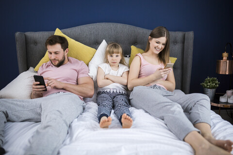 Vernachlässigtes kleines Mädchen, das mit seinen Eltern auf dem Bett sitzt und Smartphones benutzt, lizenzfreies Stockfoto