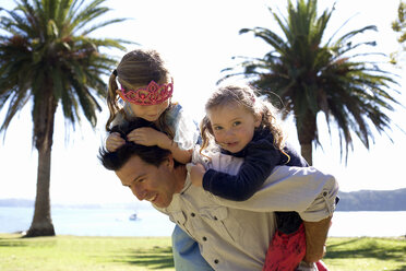 Mature man giving piggy back to daughter and friend in coastal park, New Zealand - CUF26053