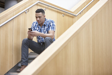 Young businessman sitting on office stairway texting on smartphone - CUF26135